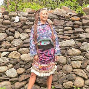 May include: A woman wearing a blue and purple patterned jacket with a white and blue patterned skirt. She is standing in front of a stone wall with green grass in the foreground.