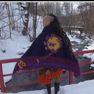 Peut inclure: Une femme portant une cape à motifs violets et bleus avec un motif de soleil jaune et un motif de papillon. Elle se tient sur un pont rouge au-dessus d'un ruisseau enneigé.