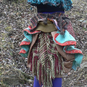 Puede incluir: Una mujer con un chaleco de crochet azul, una falda marrón y roja con flecos y botas negras. Tiene el pelo largo y castaño y está de pie en un entorno boscoso.