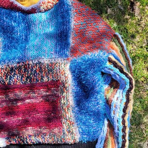 May include: A colourful patchwork sweater with a fuzzy blue texture and a black ribbed hem. The sweater is made of different knit fabrics in shades of blue, red, green, and white.