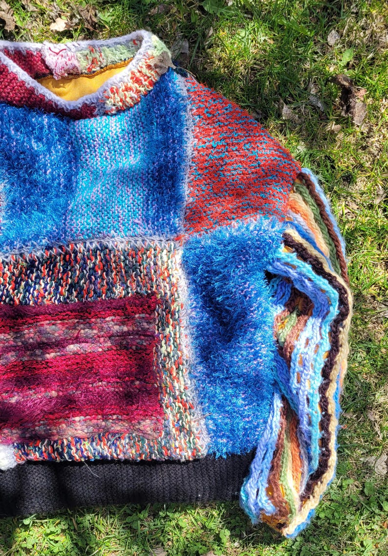 May include: A colourful patchwork sweater with a fuzzy blue texture and a black ribbed hem. The sweater is made of different knit fabrics in shades of blue, red, green, and white.