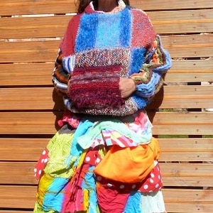 May include: A colourful patchwork sweater with a large pocket on the front. The sweater is made of different coloured yarns and has a fuzzy texture. The sweater is worn over a colourful patchwork skirt.