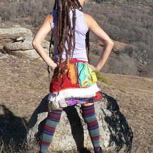 Puede incluir: Una mujer con rastas largas lleva un top morado sin mangas, una falda de retazos colorida y calcetines altos a rayas. Está de pie sobre una gran roca en un paisaje montañoso.