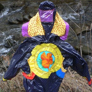 Puede incluir: Una gabardina azul con lunares blancos y parches coloridos. Los parches incluyen un círculo amarillo con una flor roja, un rectángulo morado y un patrón de estrellas amarillas.