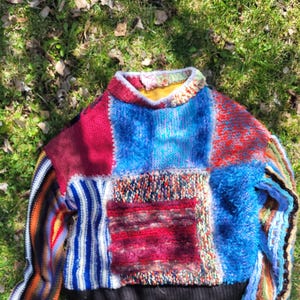 May include: A colorful patchwork sweater with a variety of textures and patterns. The sweater is made of different colored yarns, including red, blue, white, and black. The sweater has a crew neck and long sleeves.