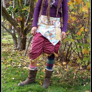 Peut inclure: Une femme portant un pull violet et une jupe en patchwork colorée avec des bottes brunes. Elle a de longues dreadlocks et se tient dans un champ de feuilles mortes.