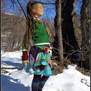 Op de afbeelding: Een vrouw met lange dreadlocks draagt een groene trui, een kleurrijke patchworkrok en gestreepte panty's. Ze staat in een besneeuwd bos met bomen op de achtergrond.