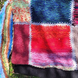 May include: A multi-coloured knitted jumper with a patchwork design. The jumper is made of different coloured yarns and has a variety of textures. The jumper is laid out on a bed of green grass.