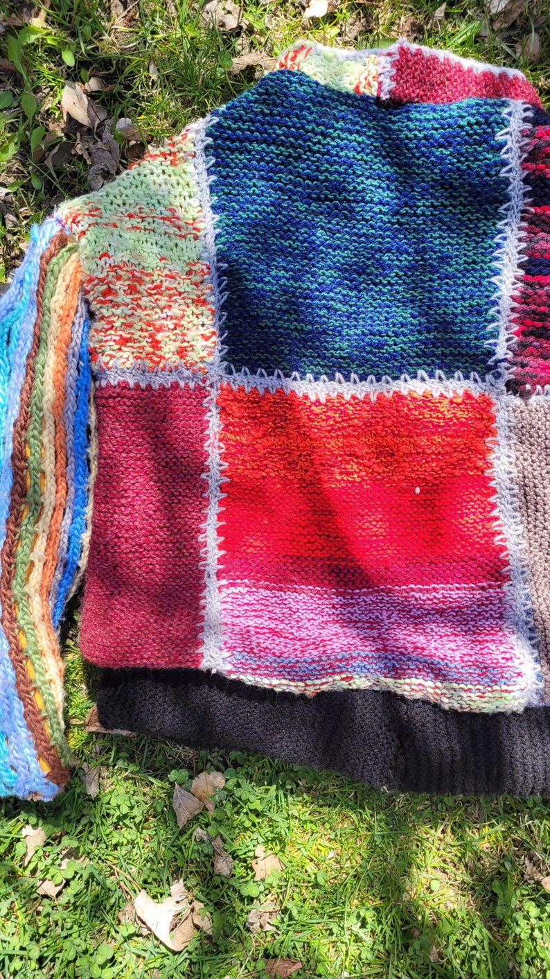May include: A multi-coloured knitted jumper with a patchwork design. The jumper is made of different coloured yarns and has a variety of textures. The jumper is laid out on a bed of green grass.