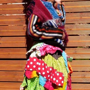 May include: A colourful patchwork skirt with a red, white and green polka dot pattern. The skirt is layered and has a ruffled hem. The person is wearing a multi-coloured striped jumper.