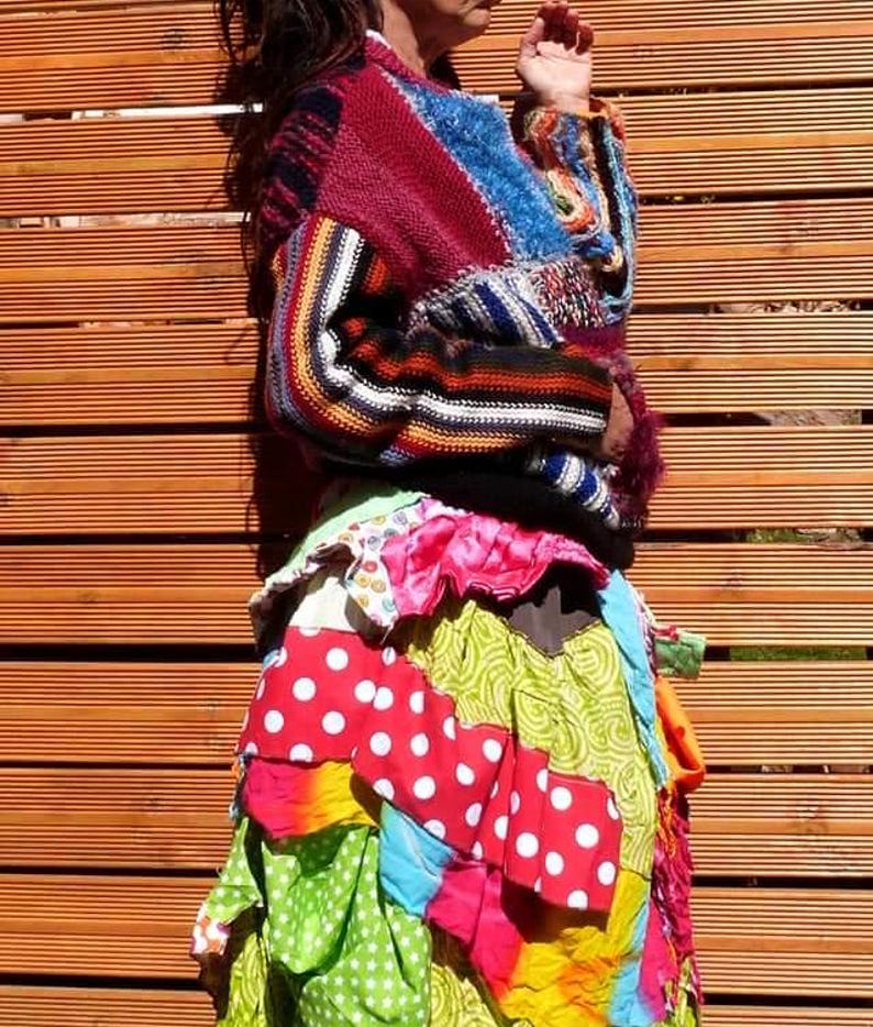 May include: A colourful patchwork skirt with a red, white and green polka dot pattern. The skirt is layered and has a ruffled hem. The person is wearing a multi-coloured striped jumper.