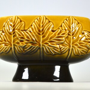 Puede incluir: Un macetero de cer&aacute;mica marr&oacute;n y amarillo con un dise&ntilde;o de hojas en relieve. El macetero tiene un cuenco ancho y redondo y una base redonda corta.