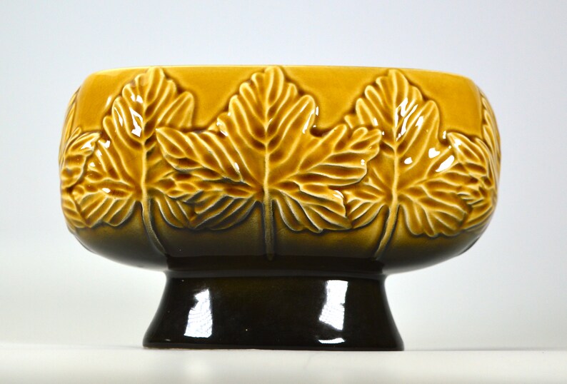 Puede incluir: Un macetero de cer&aacute;mica marr&oacute;n y amarillo con un dise&ntilde;o de hojas en relieve. El macetero tiene un cuenco ancho y redondo y una base redonda corta.