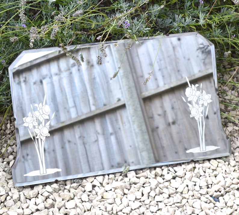 May include: A vintage beveled mirror with a decorative etched design of white flowers. The mirror has a silver frame and is sitting on a bed of white gravel.