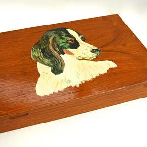 Puede incluir: Una caja de madera con una imagen pintada de la cabeza de un perro. El perro tiene pelo blanco y negro y ojos verdes. La caja es marrón y tiene una tapa con bisagras.