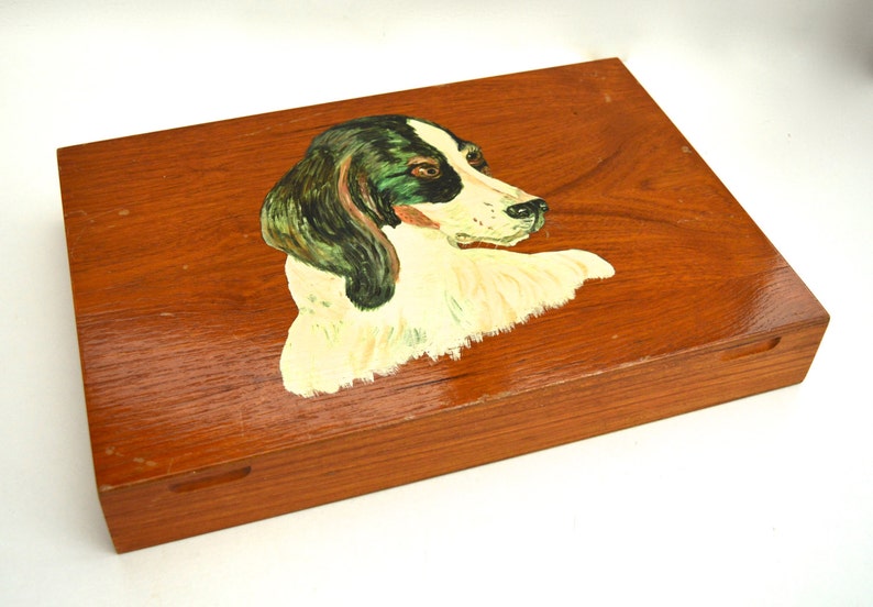 Puede incluir: Una caja de madera con una imagen pintada de la cabeza de un perro. El perro tiene pelo blanco y negro y ojos verdes. La caja es marr&oacute;n y tiene una tapa con bisagras.