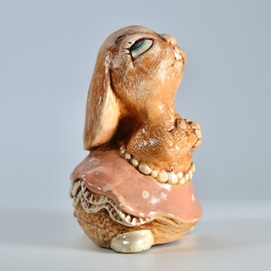 Könnte beinhalten: Eine Keramikfigur eines braunen Kaninchens, das ein rosa Kleid mit weißen Punkten und eine Perlenkette trägt. Das Kaninchen schaut nach oben und hat die Pfoten aneinander gefaltet.