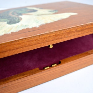 Puede incluir: Una caja de madera con tapa abatible. La parte superior de la caja est&aacute; pintada con un perro blanco con marcas marrones y verdes. El interior de la caja est&aacute; forrado con un material de terciopelo rojo oscuro.