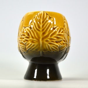 Puede incluir: Un macetero de cer&aacute;mica con un esmalte marr&oacute;n y amarillo. El macetero tiene forma de taza con una base ancha y presenta un dise&ntilde;o en relieve de hojas.