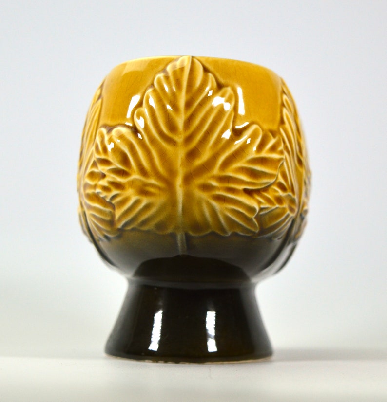 Puede incluir: Un macetero de cer&aacute;mica con un esmalte marr&oacute;n y amarillo. El macetero tiene forma de taza con una base ancha y presenta un dise&ntilde;o en relieve de hojas.