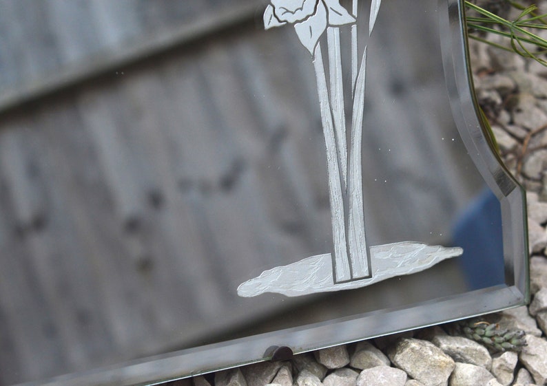 May include: A vintage beveled mirror with an etched design of a flower with long stems. The mirror has a dark gray background and a silver-colored etched design.