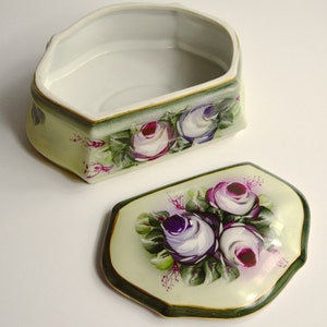 May include: An open, decorative porcelain box with a lid. The box and lid are painted with a floral design featuring purple and white roses with green leaves. The box has a light green background with a dark green border and gold trim.