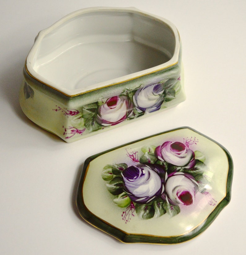 May include: An open, decorative porcelain box with a lid. The box and lid are painted with a floral design featuring purple and white roses with green leaves. The box has a light green background with a dark green border and gold trim.
