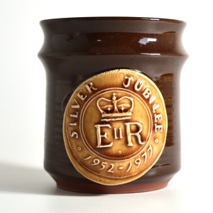 May include: A brown ceramic mug with a decorative emblem commemorating the Silver Jubilee of Queen Elizabeth II. The emblem features a crown, the initials "E II R", and the years 1952-1977. The mug has a cylindrical shape.
