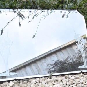 May include: A vintage beveled edge mirror with a silver frame. The mirror has an etched design of flowers on the surface. The mirror is sitting on a bed of small white rocks.