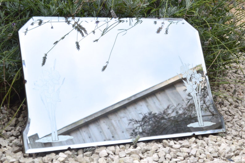 May include: A vintage beveled edge mirror with a silver frame. The mirror has an etched design of flowers on the surface. The mirror is sitting on a bed of small white rocks.