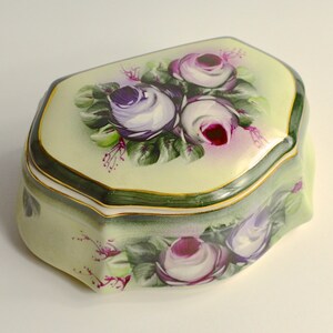 May include: A decorative, porcelain trinket box with a pale yellow background and a floral design. The box features hand-painted purple and white roses with green leaves. The edges are trimmed with gold and dark green paint.