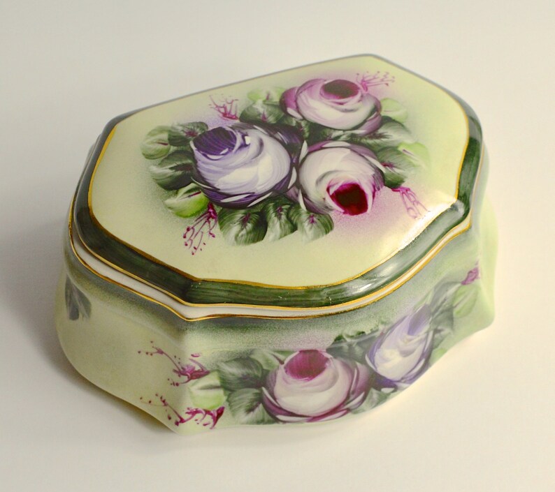 May include: A decorative, porcelain trinket box with a pale yellow background and a floral design. The box features hand-painted purple and white roses with green leaves. The edges are trimmed with gold and dark green paint.
