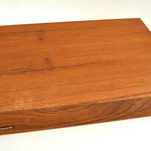 Puede incluir: Una caja de madera con tapa con bisagras y dos bisagras met&aacute;licas. La caja est&aacute; hecha de madera marr&oacute;n claro con un patr&oacute;n de veta natural.