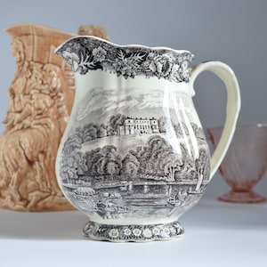May include: A white ceramic jug with a black and white landscape design. The jug has a scalloped rim and a handle. The landscape design features a house, trees, and a river.