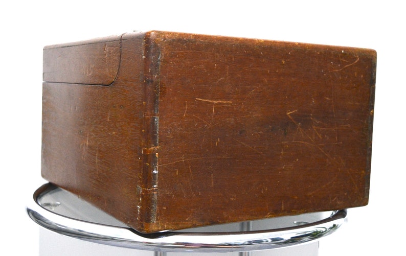 K&ouml;nnte beinhalten: Eine Holzkiste mit einem Scharnierdeckel. Die Kiste ist braun und hat eine abgenutzte, verwitterte Oberfl&auml;che. Die Kiste steht auf einem silbernen St&auml;nder.