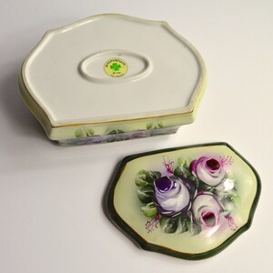 May include: A two-piece ceramic trinket box with a floral design. The lid features a bouquet of purple and pink roses on a pale yellow background, framed by a dark green border. The base is off-white with a floral pattern and a gold trim.