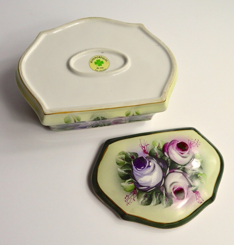 May include: A two-piece ceramic trinket box with a floral design. The lid features a bouquet of purple and pink roses on a pale yellow background, framed by a dark green border. The base is off-white with a floral pattern and a gold trim.
