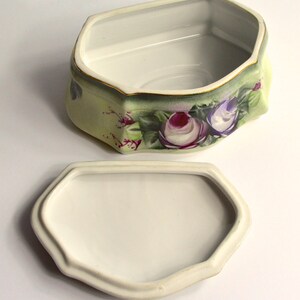 May include: A ceramic trinket box with a removable lid. The box is hexagonal with a pale green exterior, adorned with purple and white rose floral designs. The rim is trimmed with gold.