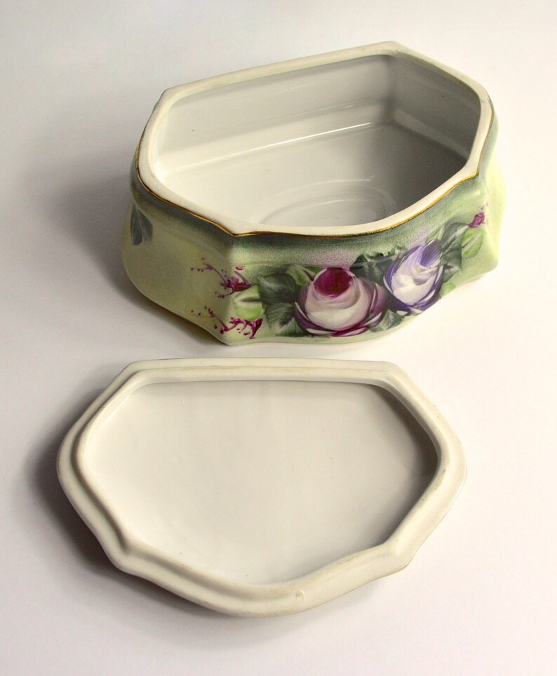 May include: A ceramic trinket box with a removable lid. The box is hexagonal with a pale green exterior, adorned with purple and white rose floral designs. The rim is trimmed with gold.