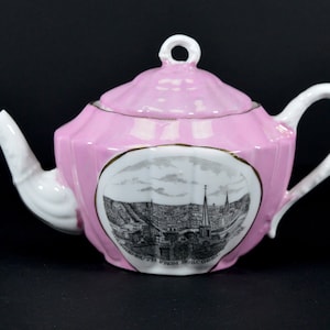 Puede incluir: Una tetera de porcelana rosa con asa y pico blancos. La tetera tiene un borde dorado y una imagen en blanco y negro de un horizonte urbano con el texto "A View of Portsmouth from Roscommon" en la parte delantera.