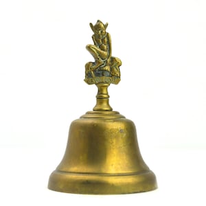 May include: A gold-coloured bell with a small figurine of a person on top. The figurine is holding a staff and is sitting on a small animal. The bell has the words "Devon Door" inscribed on it.