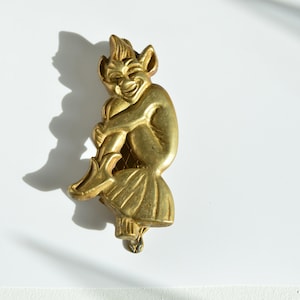Puede incluir: Un llamador de puerta de metal dorado con forma de duende sentado con una sonrisa traviesa.