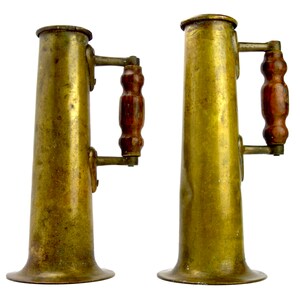 May include: Two antique brass pitchers with wooden handles and a flared base. The pitchers are both the same size and shape, and they have a simple, elegant design.