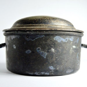 May include: A vintage silver serving dish with a lid and two black handles. The dish has a worn, aged look with dark spots and streaks.