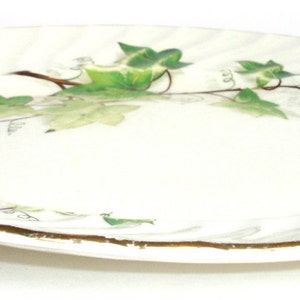 Puede incluir: Un plato de cer&aacute;mica blanco con un borde dorado y un dise&ntilde;o de hiedra verde. El plato tiene un borde festoneado y una superficie ondulada.
