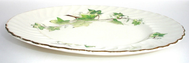 Puede incluir: Un plato de cer&aacute;mica blanco con un borde dorado y un dise&ntilde;o de hiedra verde. El plato tiene un borde festoneado y una superficie ondulada.