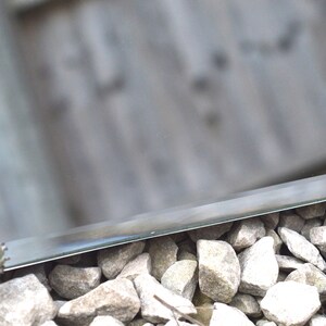 May include: A close-up of a silver metal mirror with a beveled edge, resting on a bed of small gray stones. A small green plant is visible in the reflection of the mirror.