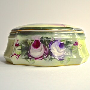 May include: A decorative ceramic box with a lid. The box is painted with a floral design featuring purple and white roses. The box has a light green background with gold trim.