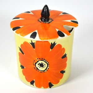 Peut inclure: Un pot à sucre en céramique avec un couvercle. Le pot et le couvercle sont décorés d'une grande fleur orange avec des accents noirs sur un fond jaune.