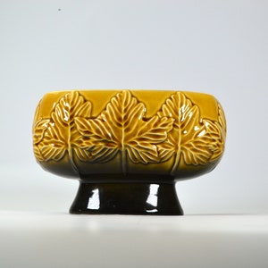 Puede incluir: Un macetero de cer&aacute;mica marr&oacute;n y amarillo con un dise&ntilde;o de hojas en relieve. El macetero tiene una base ancha y ovalada y un pie de pedestal.